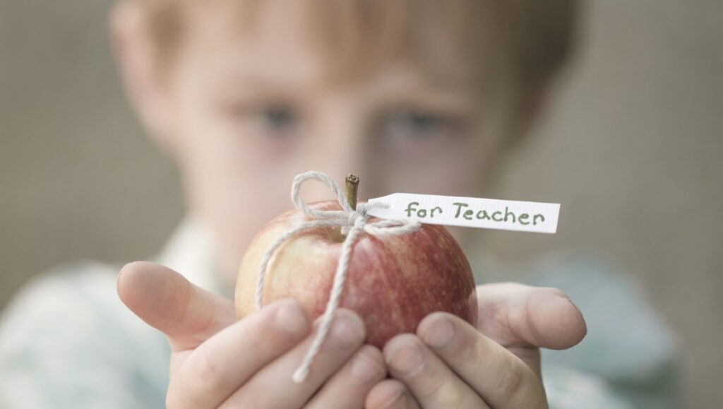Child's hands holding a small red apple tied with string and a tag reading 'for Teacher'.